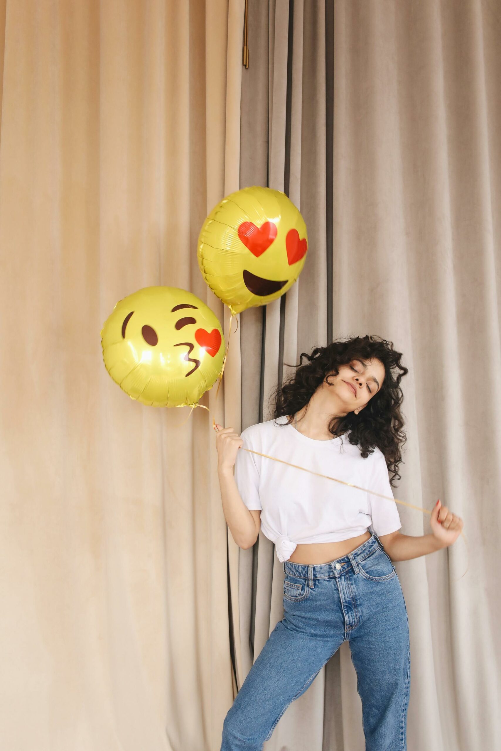 Młoda kobieta trzymająca balony z emotkami na tle zasłony – nowoczesny influencer w stylowej scenerii.